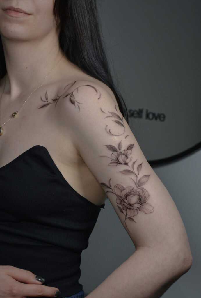 Elegancki tatuaż florystyczny fine line, rozciągający się przez ramię i górną część ręki, wykonany cienkimi liniami i delikatnym cieniowaniem. Subtelna botaniczna kompozycja w czarnym tuszu, która podkreśla kobiecą formę oraz naturalny przepływ ciała dzięki wyrafinowanemu, pełnemu gracji ruchowi. Tatuaż wykonany przez Sonyę (@inksefer) w Fineline Tattoo Gallery dla Tattoo Kraków Kolektyw Artystów.