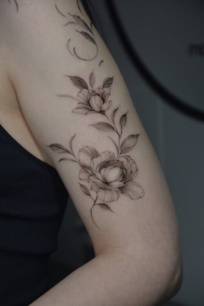 Delikatny tatuaż florystyczny fine line z motywem kwiatów i liści, umieszczony na ramieniu. Wykonany cienkimi liniami i subtelnym cieniowaniem, ten elegancki botaniczny projekt w czarnym tuszu został starannie uformowany tak, aby podążać za naturalnymi konturami ramienia, podkreślając harmonię i wyrafinowaną kobiecość. Tatuaż wykonany przez Sonyę (@inksefer) w Fineline Tattoo Gallery dla Tattoo Kraków Kolektyw Artystów.