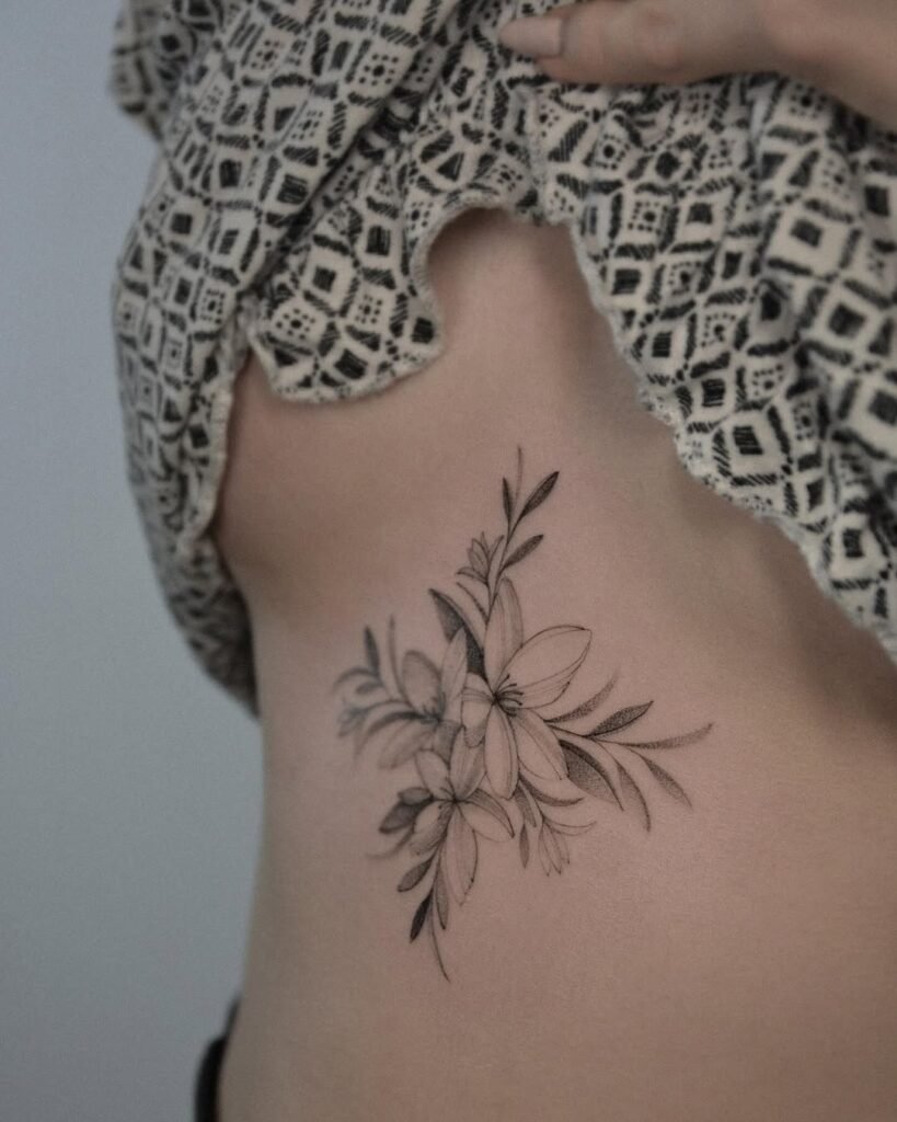 Delikatny botaniczny tatuaż fine line z warstwowymi kwiatami i smukłymi liśćmi, umieszczony na boku ciała wzdłuż żeber. Wykonany czarnym tuszem z subtelnym cieniowaniem typu dotwork, ta elegancka kompozycja podąża za naturalną krzywizną sylwetki, podkreślając lekkość, równowagę i wyrafinowaną kobiecość. Tatuaż wykonany przez Sonyę (@inksefer) w Fineline Tattoo Gallery dla Tattoo Kraków Kolektyw Artystów.