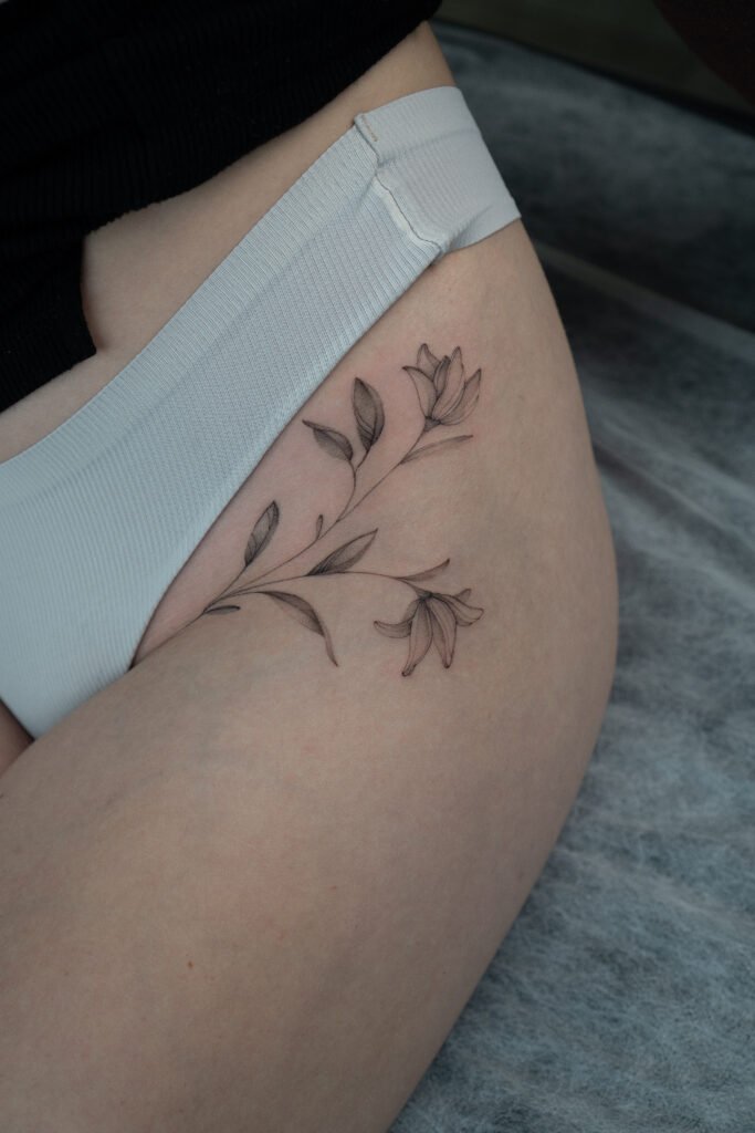 Delikatny tatuaż florystyczny fine line z motywem gałązki z kwiatami, umieszczony na udzie. Wykonany cienkimi liniami i subtelnym cieniowaniem, ten minimalistyczny botaniczny projekt w czarnym tuszu z elegancją podąża za naturalną linią ciała, podkreślając miękkość i wyrafinowaną kobiecość. Tatuaż wykonany przez Sonyę (@inksefer) w Fineline Tattoo Gallery dla Tattoo Kraków Kolektyw Artystów.