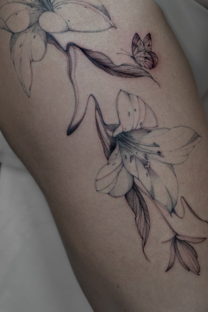 Delikatny tatuaż fine line z motywem lilii i małego motyla, wykonany cienkimi liniami i subtelnym cieniowaniem typu dotwork. Elegancka botaniczna kompozycja w czarnym tuszu, podkreślająca lekkość, naturalny ruch oraz wyrafinowany detal. Tatuaż wykonany przez Sonyę (@inksefer) w Fineline Tattoo Gallery dla Tattoo Kraków Kolektyw Artystów.