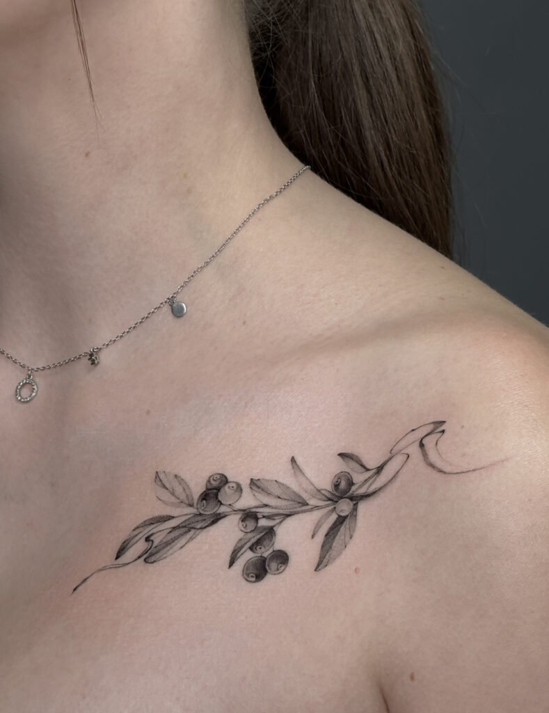 Delikatny tatuaż fine line przedstawiający gałązkę oliwną z liśćmi i drobnymi owocami, umieszczony na obojczyku. Subtelny, minimalistyczny botaniczny projekt wykonany czarnym tuszem, stworzony z precyzyjnych, cienkich linii — idealny dla osób ceniących eleganckie i kobiece tatuaże. Tatuaż wykonany przez Sonyę (@inksefer) w Fineline Tattoo Gallery dla Tattoo Kraków Kolektyw Artystów.