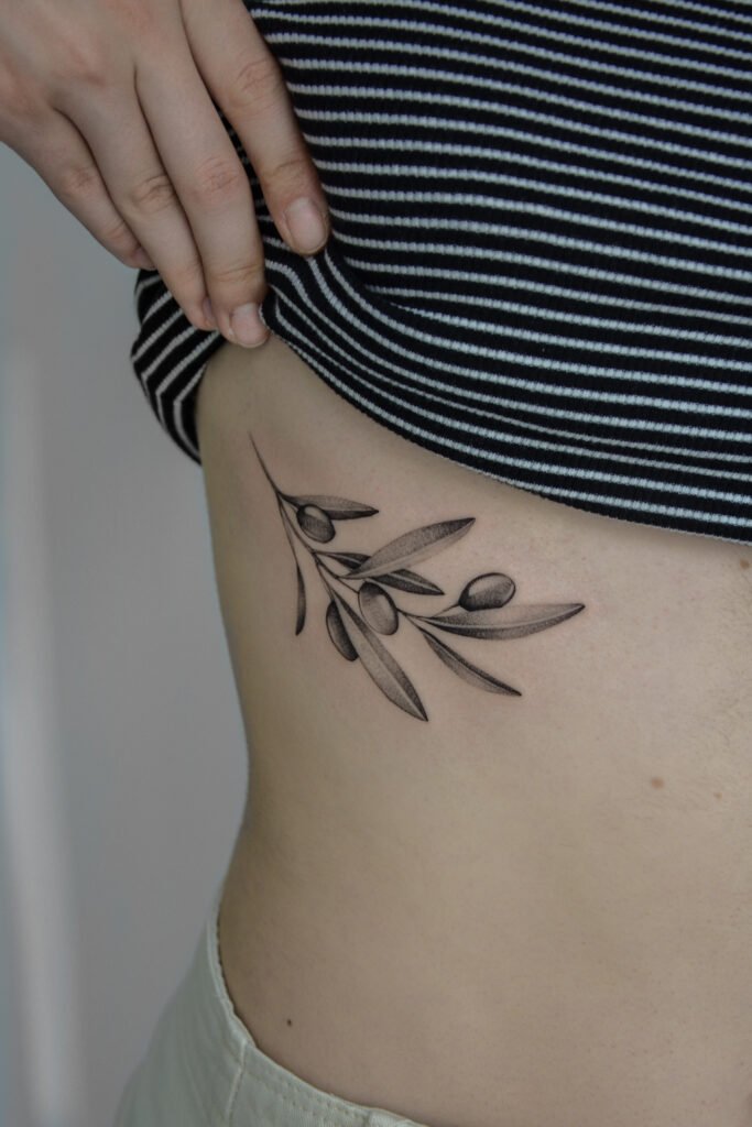 Delikatny tatuaż fine line z motywem gałązki oliwnej z owocami, umieszczony na boku ciała. Wykonany cienkimi liniami i subtelnym cieniowaniem typu dotwork, ten minimalistyczny botaniczny projekt w czarnym tuszu z elegancją i powściągliwością podkreśla naturalną linię sylwetki. Tatuaż wykonany przez Sonyę (@inksefer) w Fineline Tattoo Gallery dla Tattoo Kraków Kolektyw Artystów.