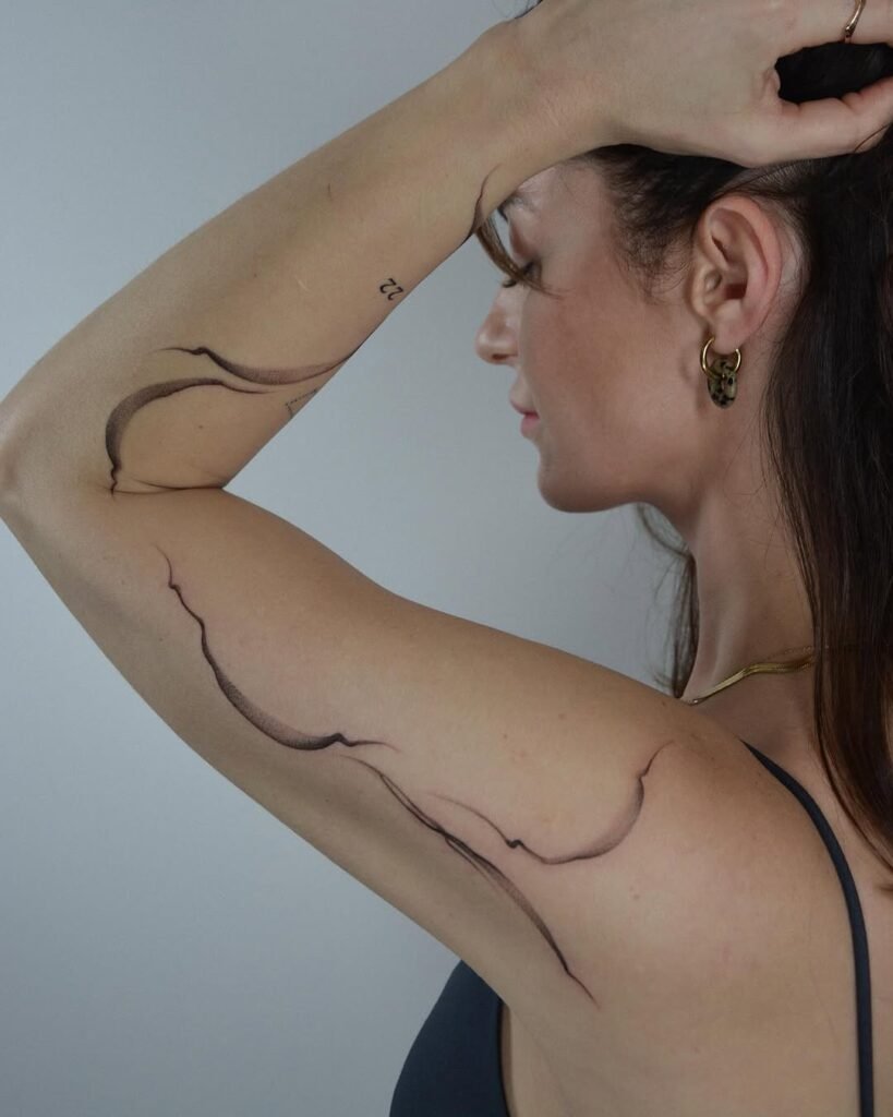 Minimalistyczny tatuaż fine line typu body-flow, złożony z abstrakcyjnych, organicznych linii rozciągających się na górnej części ramienia oraz po jego wewnętrznej stronie. Wykonany czarnym tuszem z miękkim, tonalnym cieniowaniem, ten współczesny projekt podąża za ruchem mięśni i naturalną anatomią ciała, tworząc płynny, elegancki i nowoczesny rytm wizualny. Tatuaż wykonany przez Sonyę (@inksefer) w Fineline Tattoo Gallery dla Tattoo Kraków Kolektyw Artystów.