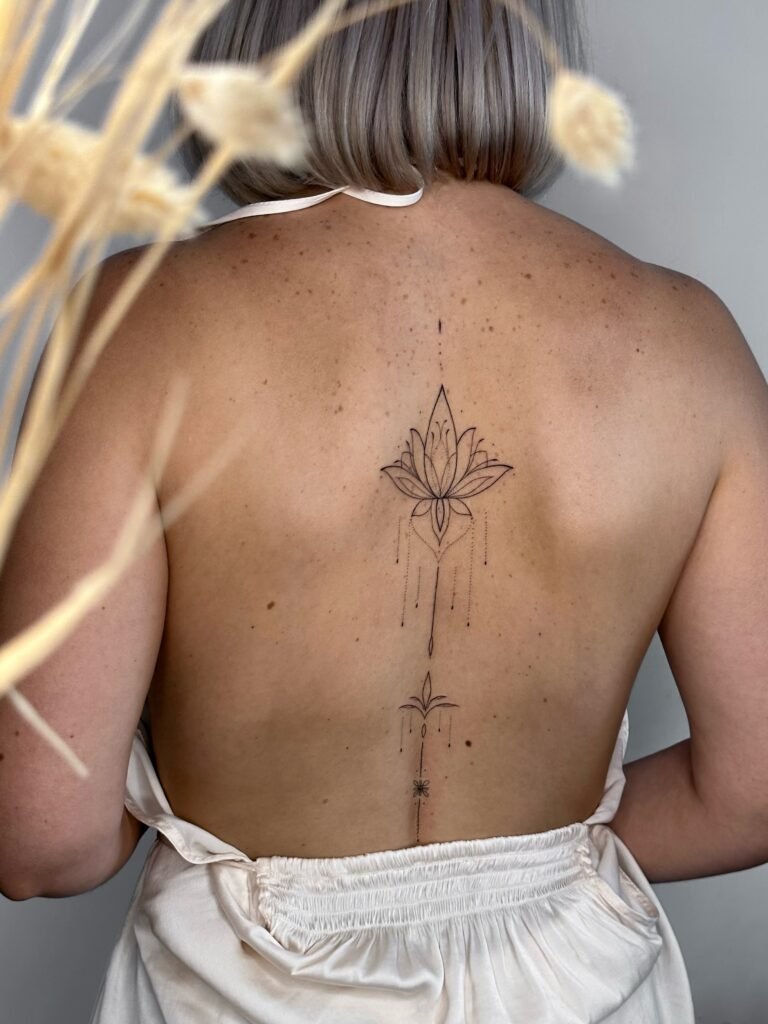Ornamentalny tatuaż fine line z motywem lotosu, biegnący wzdłuż kręgosłupa. Delikatny, kobiecy projekt wykonany czarnym tuszem, łączący elementy botaniczne i geometryczne z harmonijnym, pionowym układem. Tatuaż wykonany przez Lorę (@lora.artss) w Fineline Tattoo Gallery w Krakowie dla Tattoo Krakow Kolektyw Artystów.