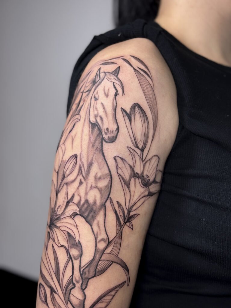 Szczegółowy tatuaż na górnej części ramienia, przedstawiający konia otoczonego liliami i liśćmi w wyrafinowanej kompozycji botanicznej. Elegancki projekt inspirowany naturą, łączący ekspresyjną kreskę z motywami florystycznymi, balansujący pomiędzy siłą a delikatnością. Tatuaż wykonany przez Olgę Sankovą (@olga_sankova_tattoo) w Fineline Tattoo Gallery dla Tattoo Kraków Kolektyw Artystów.