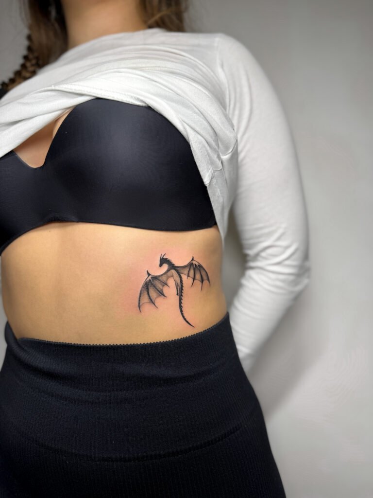 Minimalistyczny tatuaż przedstawiający smoka, wykonany czarnym tuszem i umieszczony na boku żeber. Czysty, a zarazem wyrazisty projekt inspirowany fantastyką, podkreślający siłę, wolność i indywidualność poprzez prostą, ekspresyjną sylwetkę. Tatuaż wykonany przez Olgę Sankovą (@olga_sankova_tattoo) w Fineline Tattoo Gallery dla Tattoo Kraków Kolektyw Artystów.