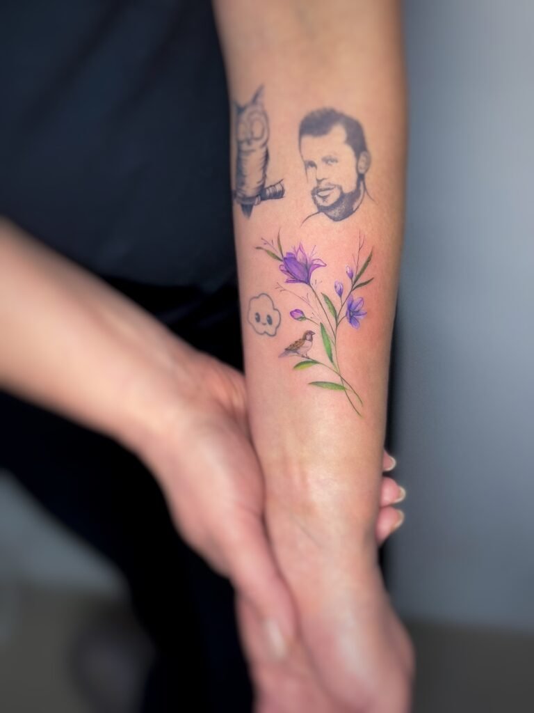 Intensywny tatuaż przedstawiający fioletowy kwiat, umieszczony wśród istniejących tatuaży na przedramieniu. Niewielki, lecz wyrazisty projekt kolorystyczny, który dodaje żywego botanicznego akcentu i naturalnie komponuje się z otaczającymi tatuażami. Tatuaż wykonany przez Olgę Sankovą (@olga_sankova_tattoo) w Fineline Tattoo Gallery dla Tattoo Kraków Kolektyw Artystów.