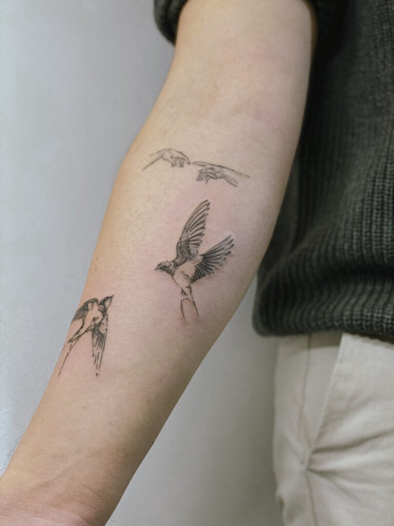 Minimalistyczna kompozycja tatuażu fine line na przedramieniu, przedstawiająca lecące ptaki oraz delikatne dłonie inspirowane sztuką klasyczną. Wykonana z subtelnym cieniowaniem i precyzyjną kreską, tworzy lekki, symboliczny i zwiewny projekt. Tatuaż wykonany przez Yarę (@yaroslavaa.tattoo) w Fineline Tattoo Gallery dla Tattoo Kraków Kolektyw Artystów.