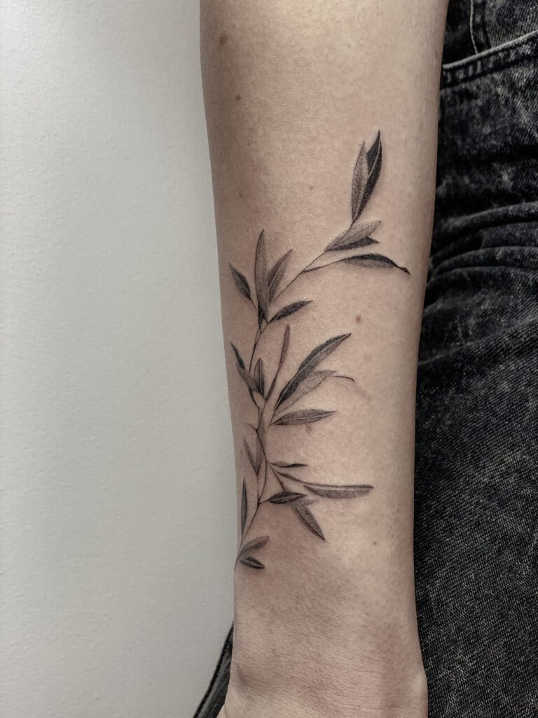 Minimalistyczny botaniczny tatuaż fine line z motywem delikatnej, liściastej gałązki, wykonany cienkimi liniami i miękkim czarnym cieniowaniem. Kompozycja podąża za naturalnym kształtem przedramienia, nadając projektowi lekki i elegancki przepływ. Tatuaż wykonany przez Yarę (@yaroslavaa.tattoo) w Fineline Tattoo Gallery dla Tattoo Kraków Kolektyw Artystów.