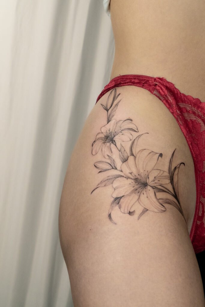 Delikatny, wielkoformatowy tatuaż kwiatowy na biodrze przedstawiający lilie. Praca wykonana przez Olgę Sankovą (@olga_sankova_tattoo) w Fineline Tattoo Gallery dla Tattoo Krakow Artists Collective. Kompozycja wykorzystuje technikę fine line oraz miękkie cieniowanie, a jej projekt został stworzony tak, aby harmonijnie współgrać z naturalnymi krzywiznami kobiecego ciała.